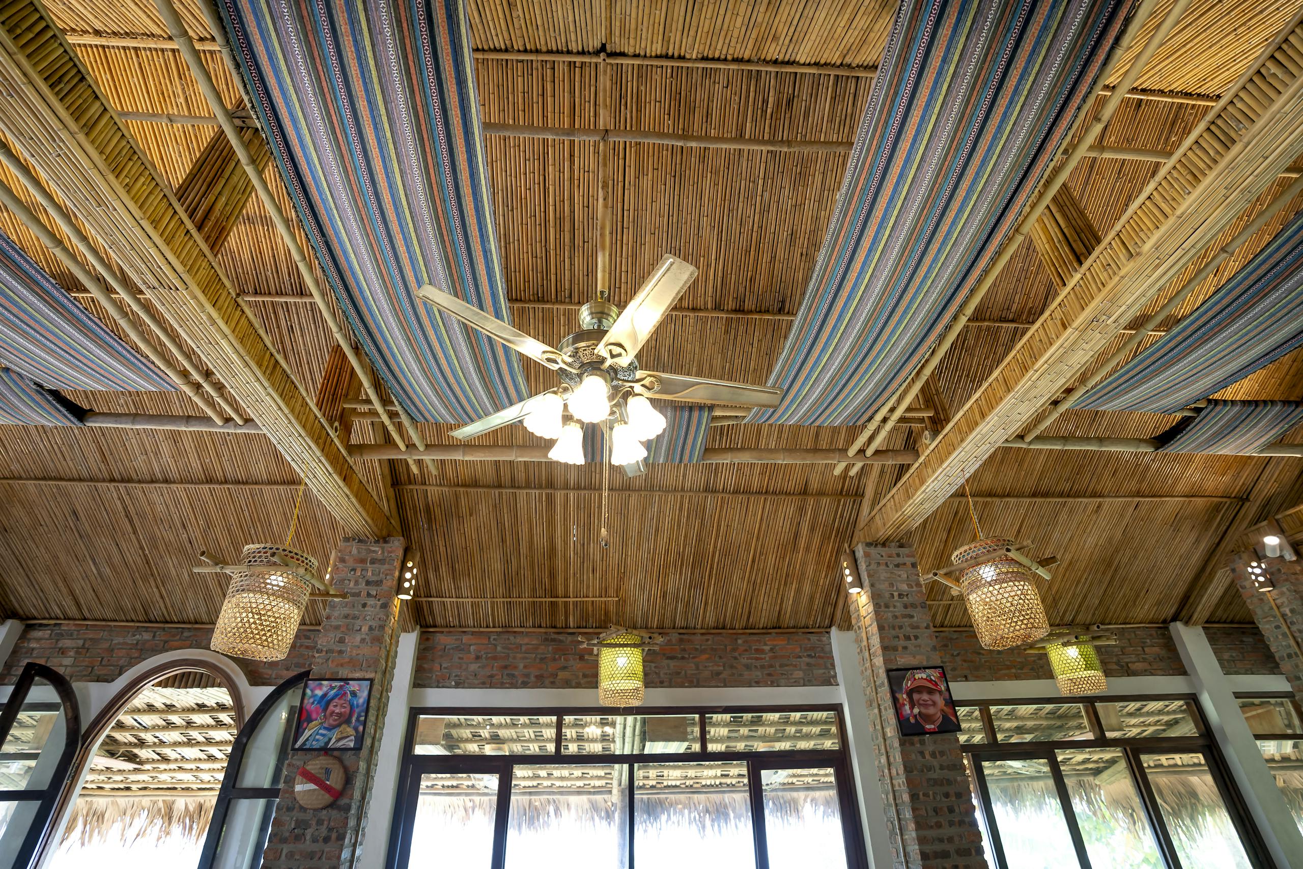 From below of glowing chandelier hanging on wooden roof with traditional colorful fabric near illuminated lamps in tropical cafeteria with windows